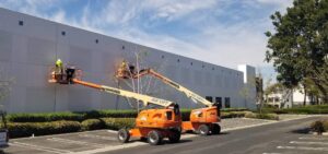Two workers on orange JLG lifts performing maintenance on a large white building